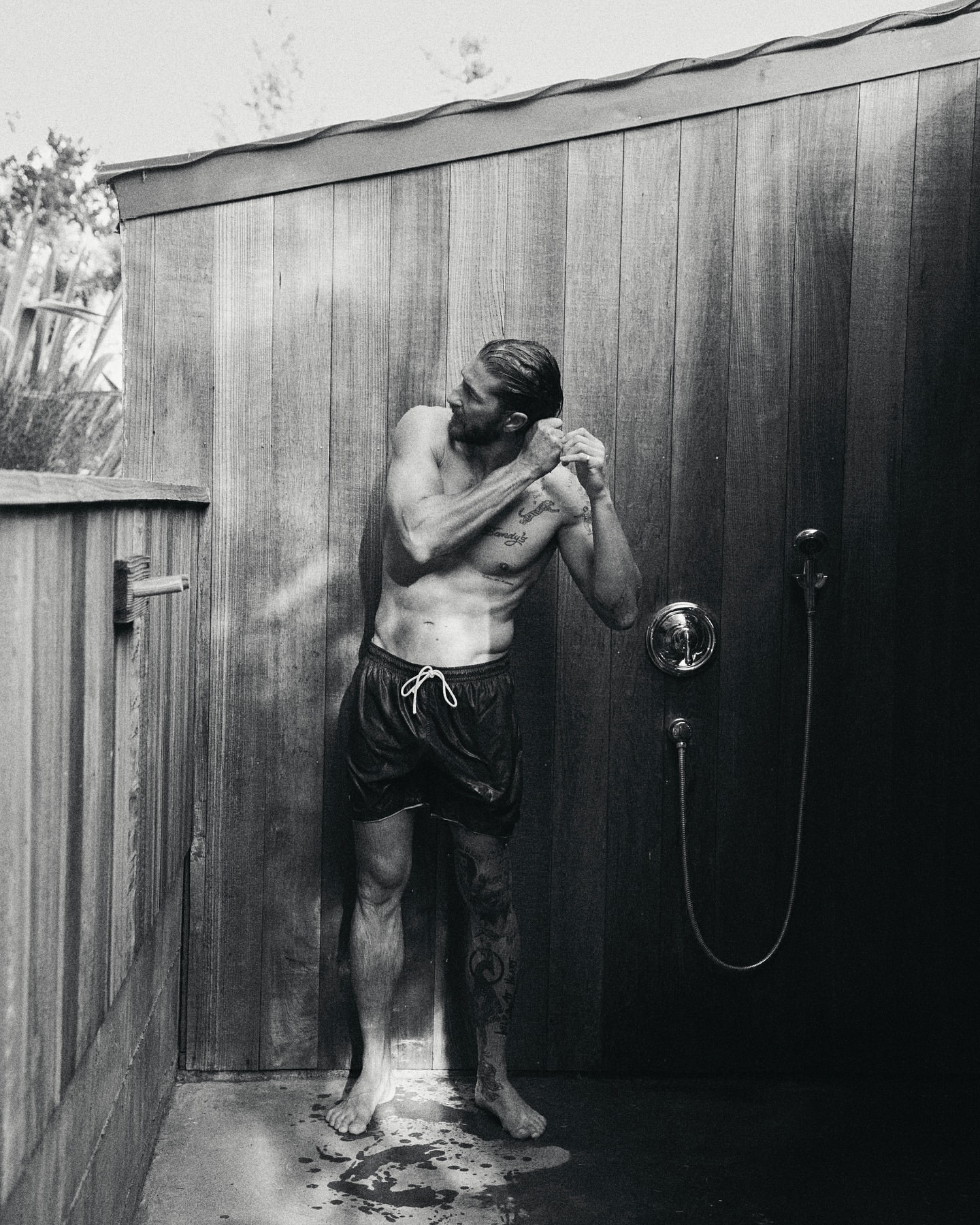 Monochrome outdoor shot of a muscular, shirtless adult male in swim trunks standing under a shower with a shower head in a wood stall.