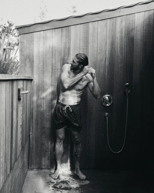 Monochrome outdoor shot of a muscular, shirtless adult male in swim trunks standing under a shower with a shower head in a wood stall.