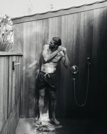 Monochrome outdoor shot of a muscular, shirtless adult male in swim trunks standing under a shower with a shower head in a wood stall.