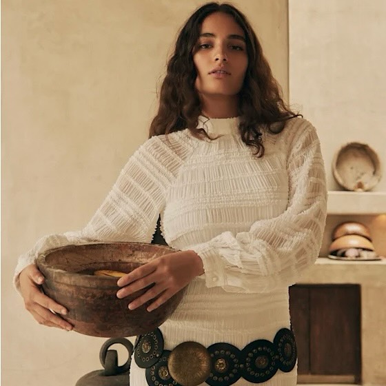 Brunette woman in white dress holds a wooden bowl in an indoor setting with neutral background, showcasing fashion and visual texture.