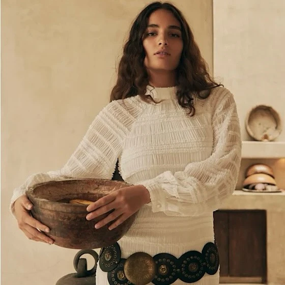 Brunette woman in white dress holds a wooden bowl in an indoor setting with neutral background, showcasing fashion and visual texture.
