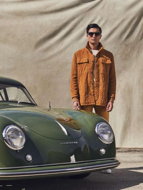 A stylish man in a brown jacket poses next to a classic green Porsche in front of a neutral backdrop, creating a vintage automotive fashion shot.