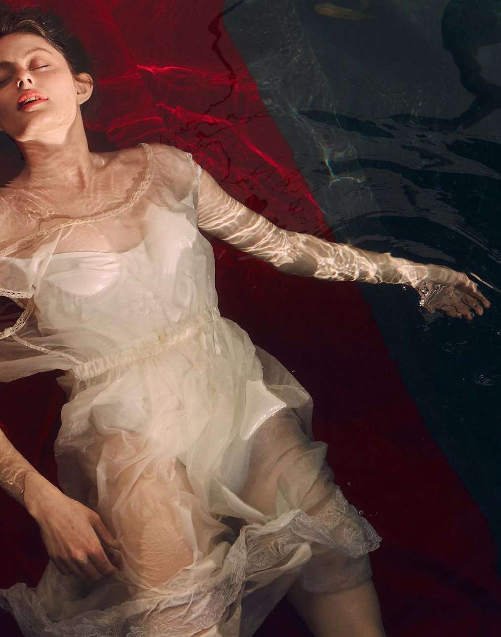 A woman floats in dark water, wearing a vintage-style white dress. Red and black colors contrast in the water around her.