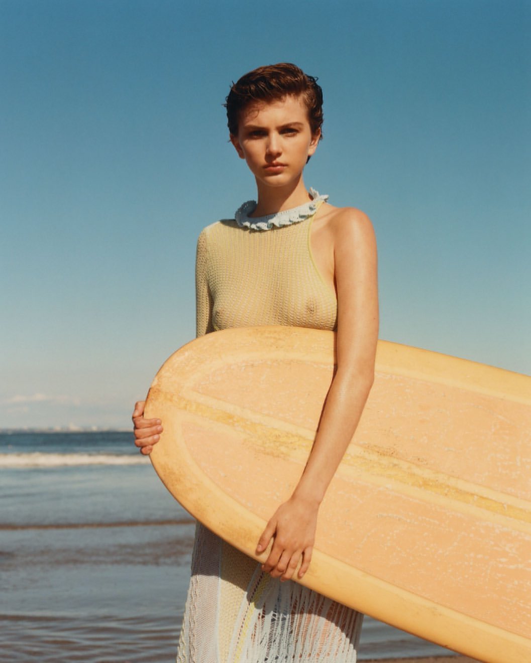 Woman on beach holds a surfboard wearing a light-green, one-shoulder knit top and light-colored fringe skirt in a portrait shot.
