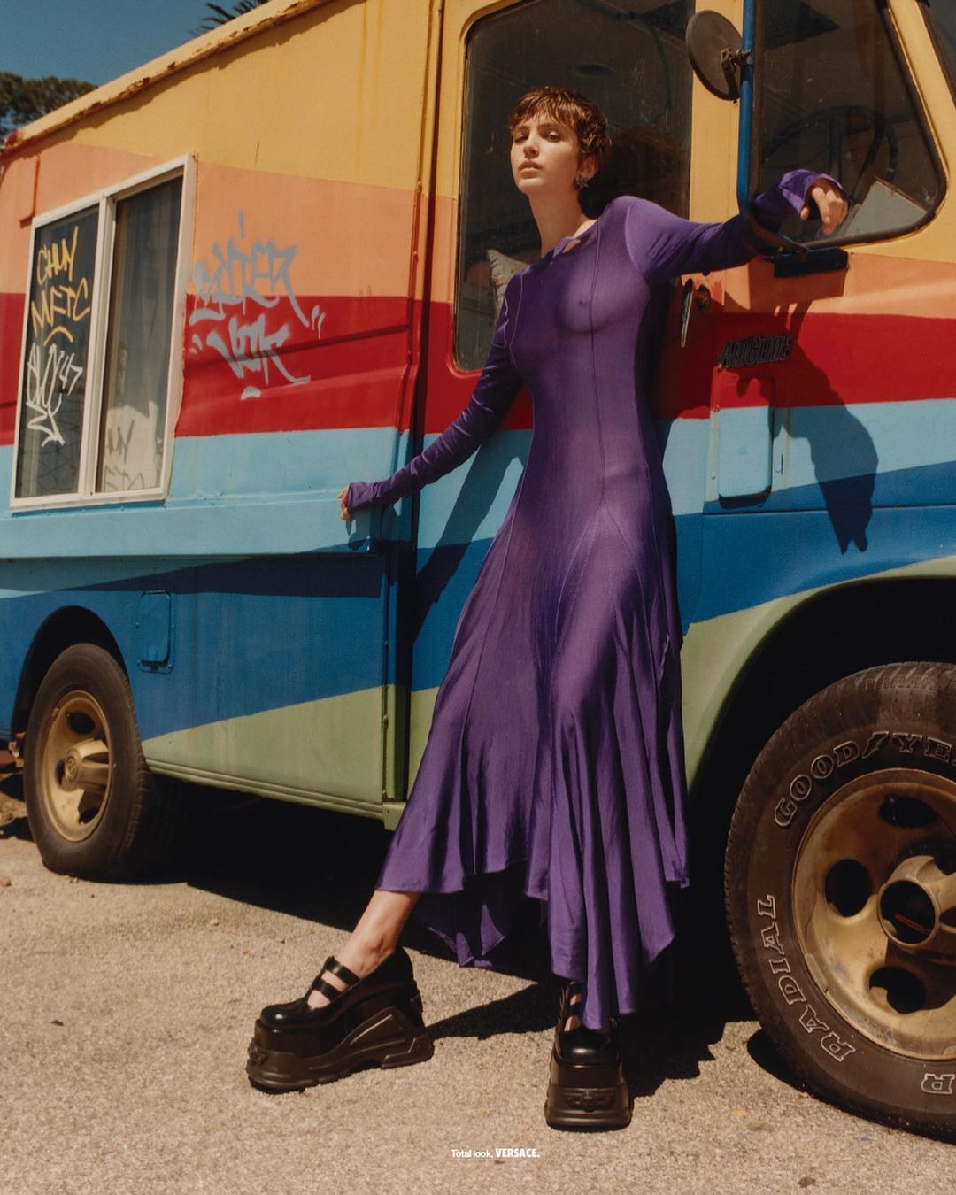 Model in a long purple dress and platform shoes leaning against a vibrant multi-colored food truck outdoors