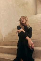 Woman with long blonde hair wearing a black long-sleeve dress seated on concrete stairs.
