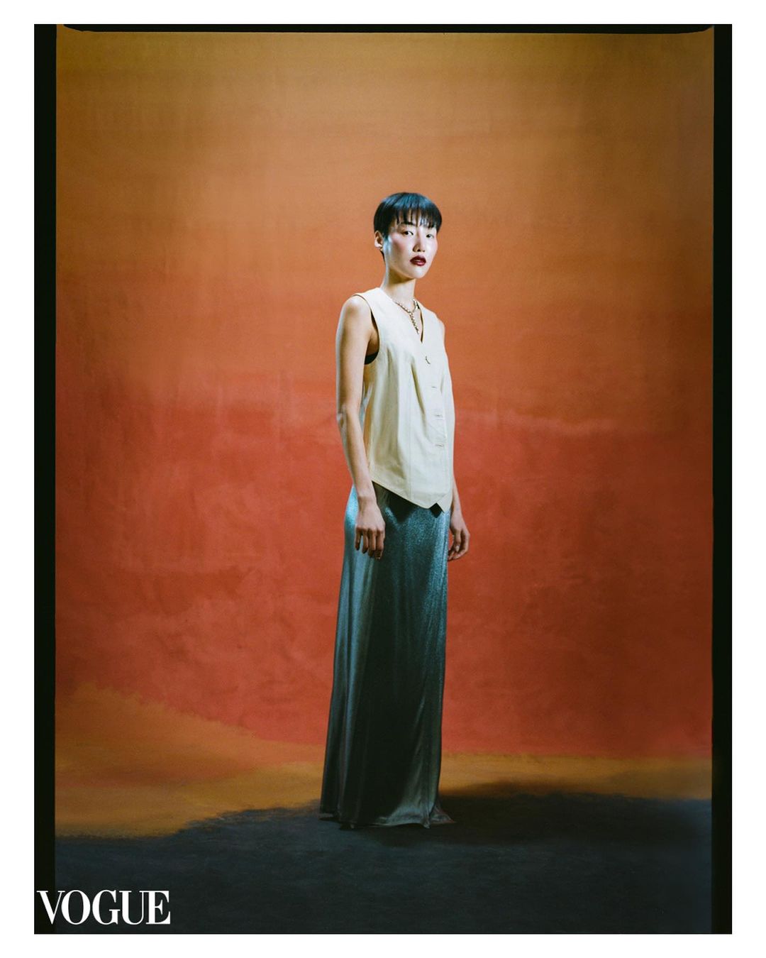 A short-haired female model poses in a sleeveless top and long skirt against an orange backdrop, illuminated by studio lighting.
