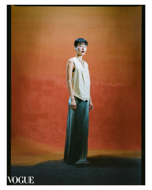A short-haired female model poses in a sleeveless top and long skirt against an orange backdrop, illuminated by studio lighting.