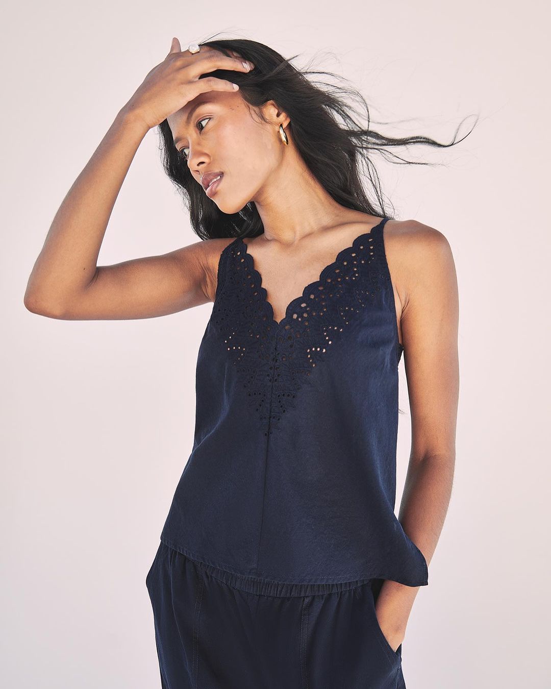 A woman models a sleeveless, dark-blue, V-neck top and matching pants against a neutral background in a fashion or beauty context.