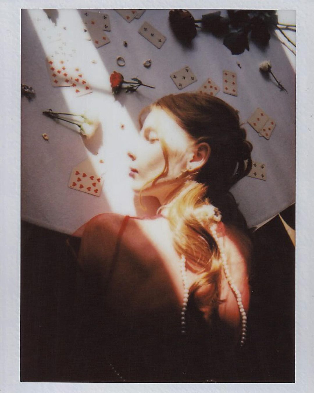 Artistic portrait of a woman lying among playing cards and roses, with soft lighting and a dreamlike aesthetic in a polaroid-style frame.
