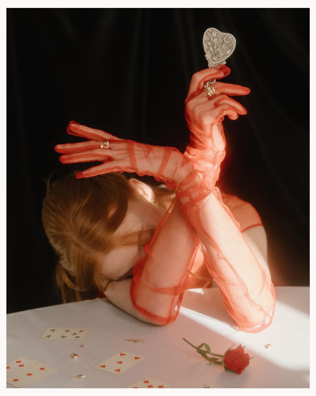 Woman's hands in red gloves holding a heart mirror among playing cards and a rose on a white table against a black background.