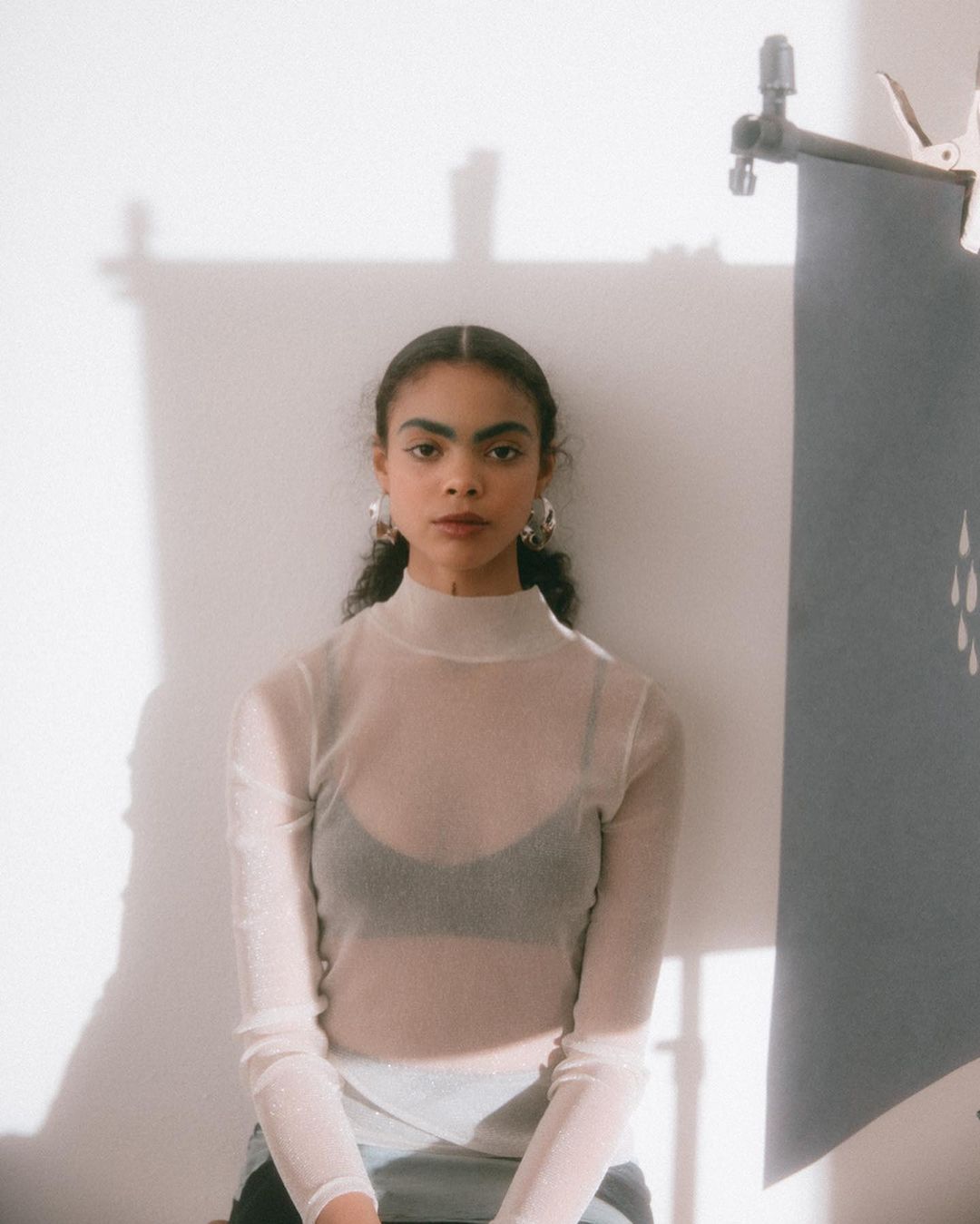 A young woman in a white top poses in a brightly lit room with a grey banner nearby, showcasing a simple yet elegant composition.