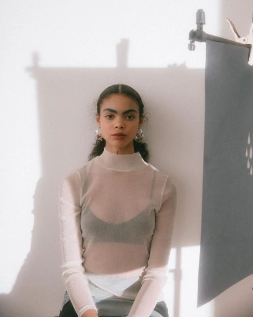 A young woman in a white top poses in a brightly lit room with a grey banner nearby, showcasing a simple yet elegant composition.