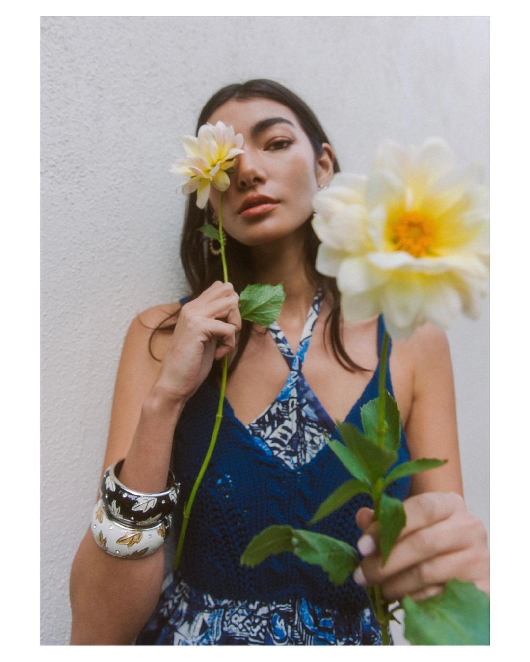 Portrait of a young woman holding white dahlia flowers to her face, wearing a blue patterned dress, and standing against a textured wall.