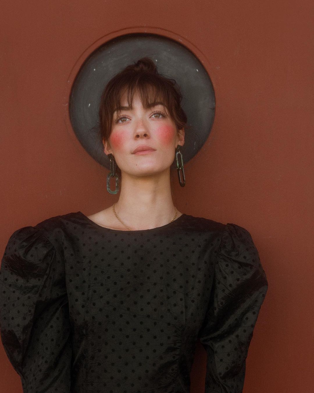 Portrait of a woman with updo, wearing earrings and a black patterned blouse, posing against a reddish-brown wall with a circular window.