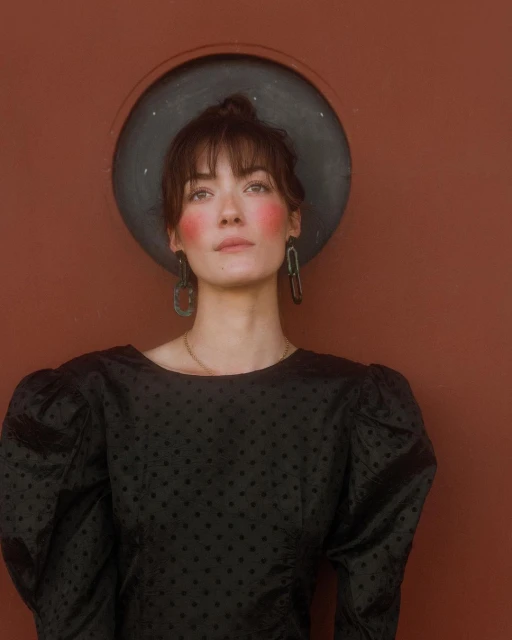 Portrait of a woman with updo, wearing earrings and a black patterned blouse, posing against a reddish-brown wall with a circular window.
