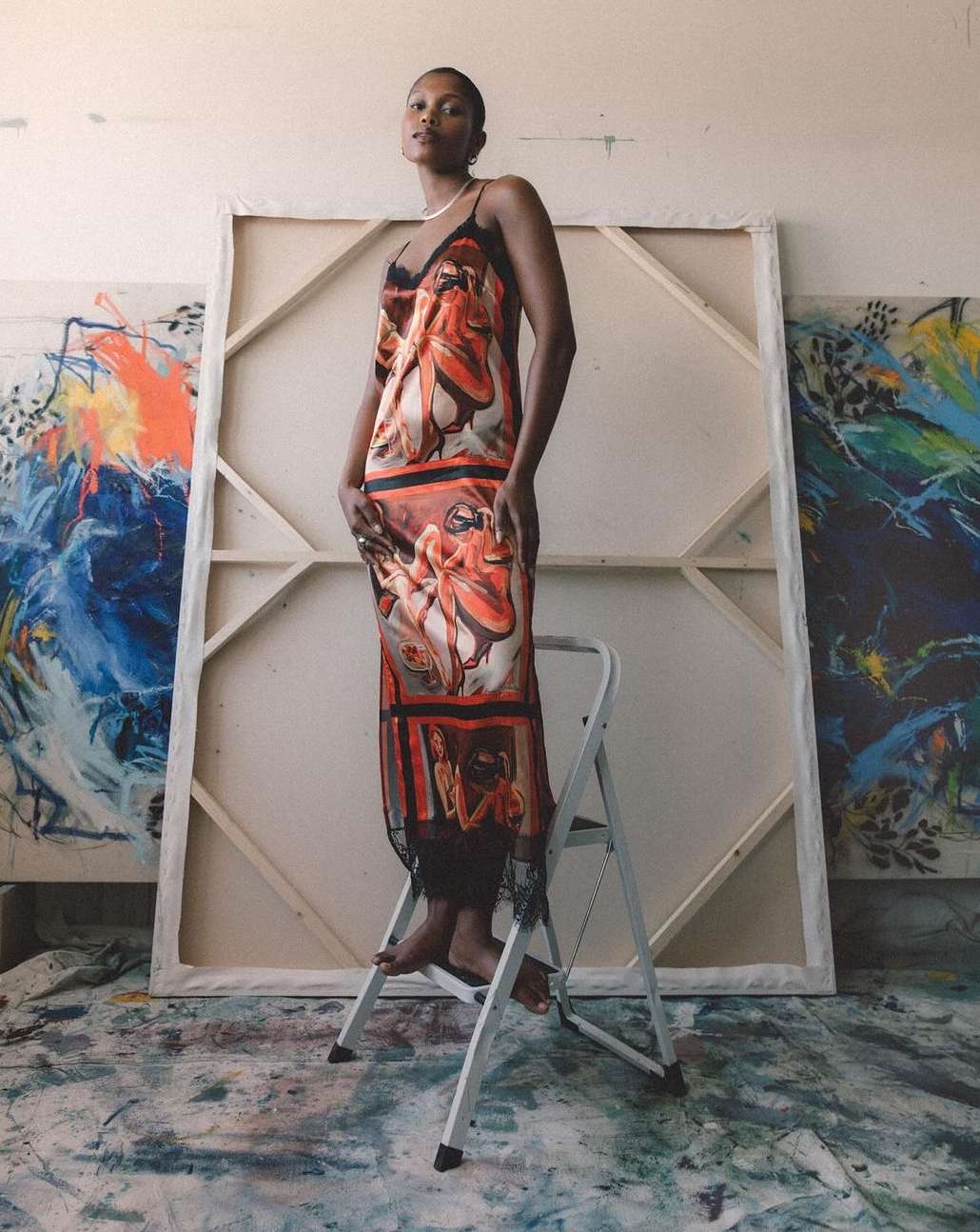 Woman in patterned dress standing on a step stool in an artist's studio with painted canvases in the background, natural light.