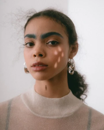 Portrait of a young woman with dark curly hair, blue eyebrows, and large silver hoop earrings wearing a sheer neutral top against a white wall.
