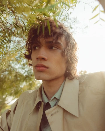 Close-up portrait of a young man with curly brown hair wearing a tan trench coat under green foliage with warm sunlight.
