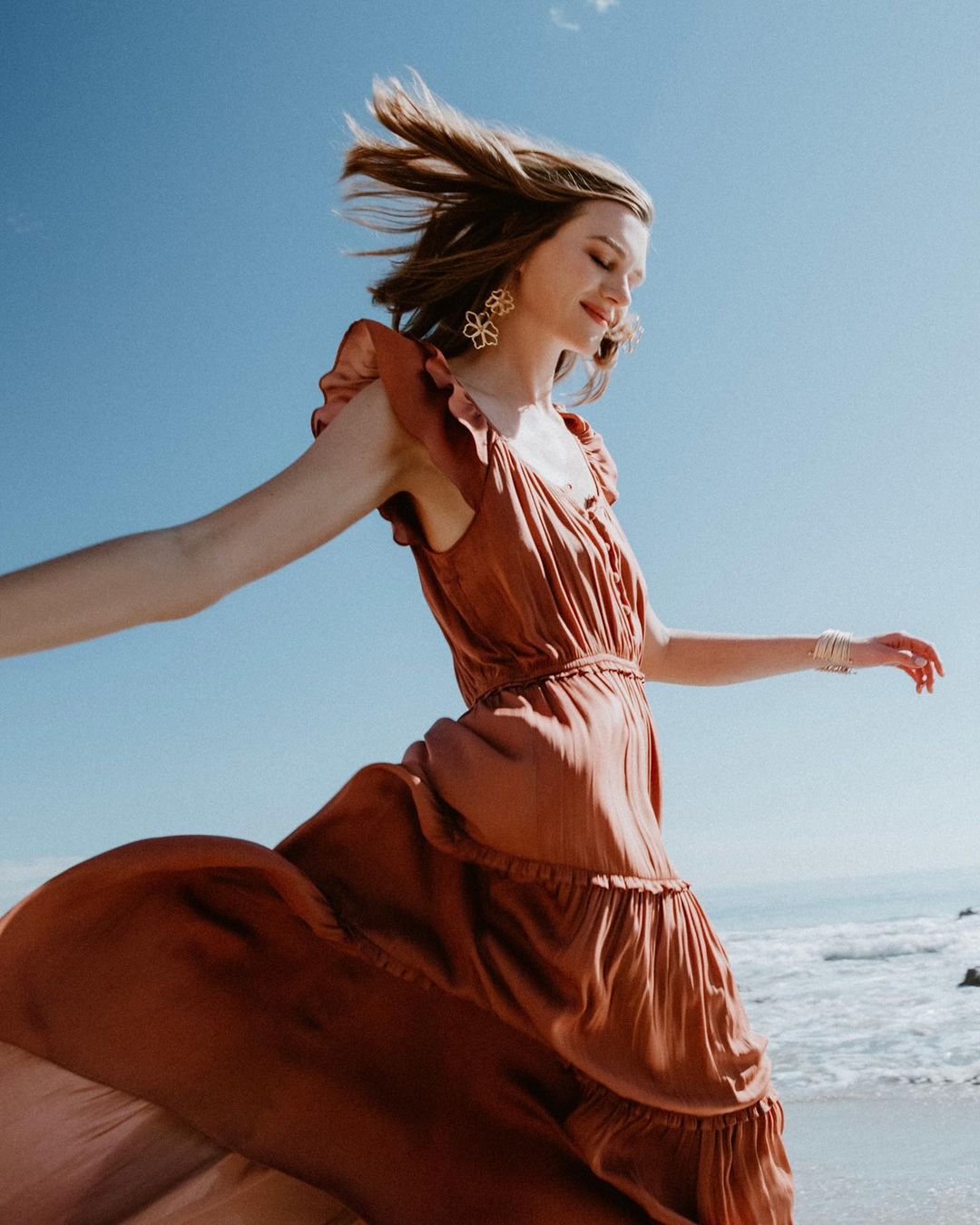 Woman in a long terracotta dress walking on the beach with blue sky. The dress flows in the breeze and she wears flower earrings.