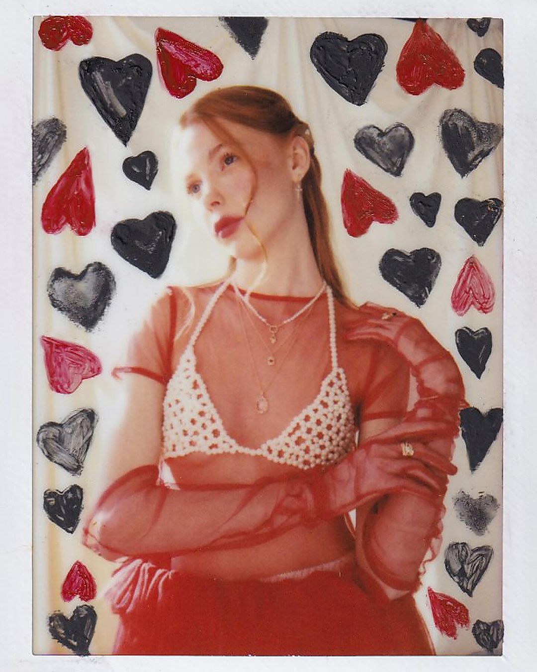 Portrait of a young woman in a sheer red outfit and pearl jewelry, posing in front of a background of painted red and black hearts.