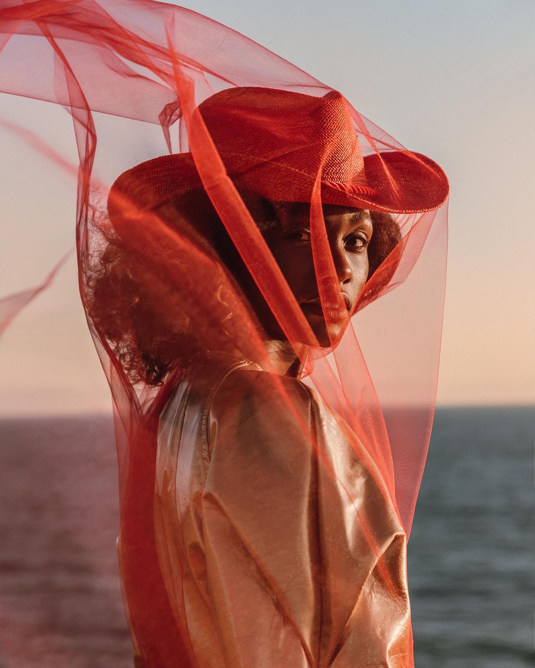 Elegant portrait of a Black woman wearing a red hat and a sheer red veil with an ocean and sky background at golden hour.