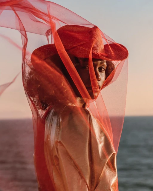 Elegant portrait of a Black woman wearing a red hat and a sheer red veil with an ocean and sky background at golden hour.