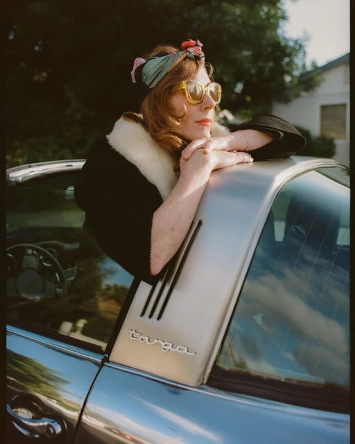 A woman wearing a turban and sunglasses leans on the silver back panel of a Porsche Targa in an outdoor setting on a sunny day.