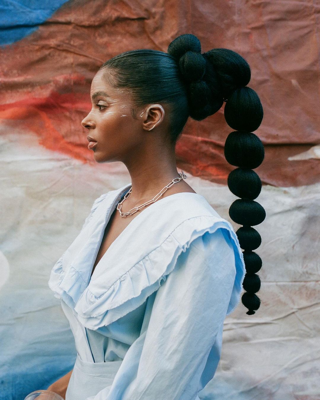 Profile portrait of a Black woman with a creative hairstyle, wearing a light blue dress, against a painterly backdrop in soft light.