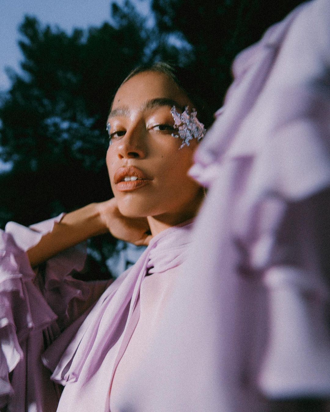 Close-up of a woman with decorative floral accents on her face, wearing a lilac-colored ruffled blouse, with a dark, leafy background.