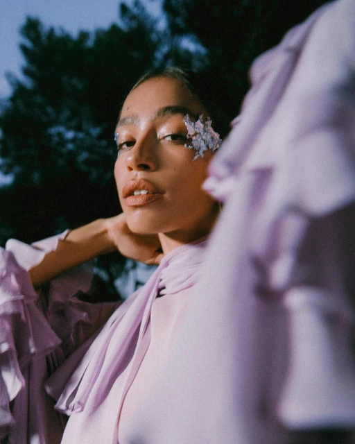 Close-up of a woman with decorative floral accents on her face, wearing a lilac-colored ruffled blouse, with a dark, leafy background.