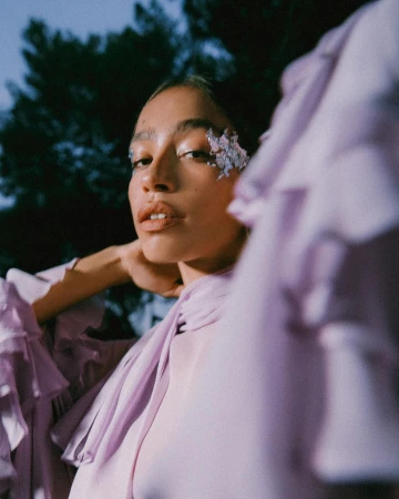 Close-up of a woman with decorative floral accents on her face, wearing a lilac-colored ruffled blouse, with a dark, leafy background.