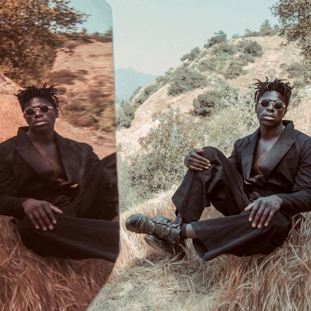 A Black male model in a black suit and round sunglasses sits on a grassy hill with a backdrop of mountains and trees in soft lighting.