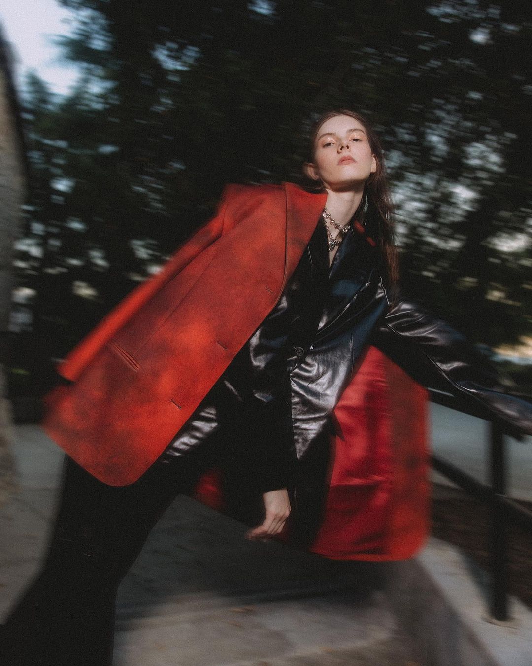 Fashionable woman in a red blazer poses outdoors with blurred motion, showcasing an edgy look with a black top and dark pants.