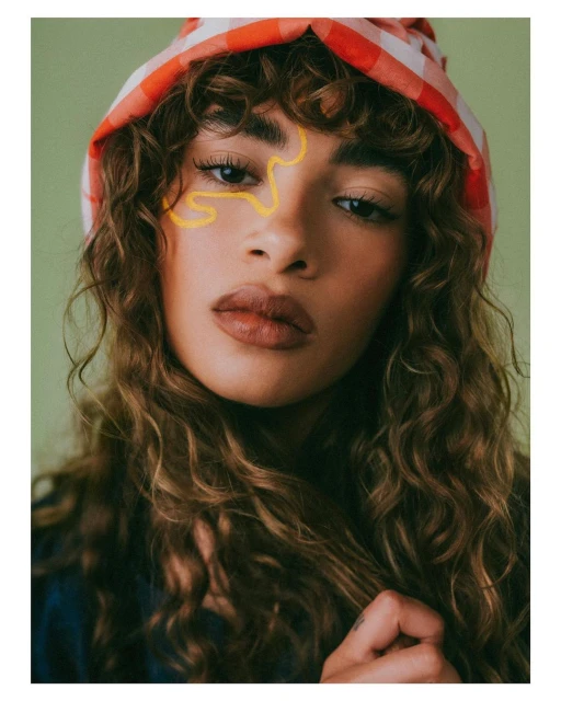 Close-up portrait of a woman with wavy hair wearing a red and white headscarf, featuring a yellow artistic line on her face, indoors.