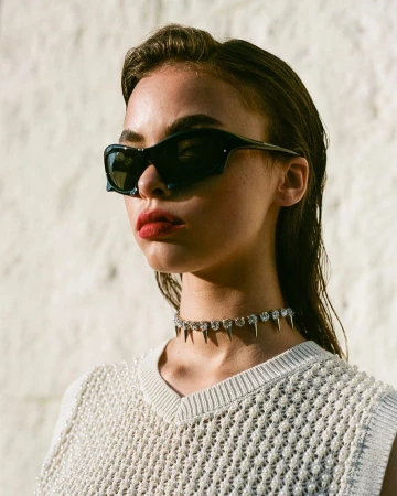 Close-up portrait of a woman wearing black wrap sunglasses, a spiked crystal choker, and a white beaded sweater.