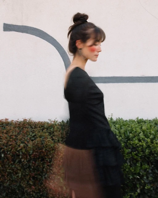 Side-view studio portrait of a brunette woman with a bun, in a black top and reddish blush standing outdoors near a light wall and green shrubs.