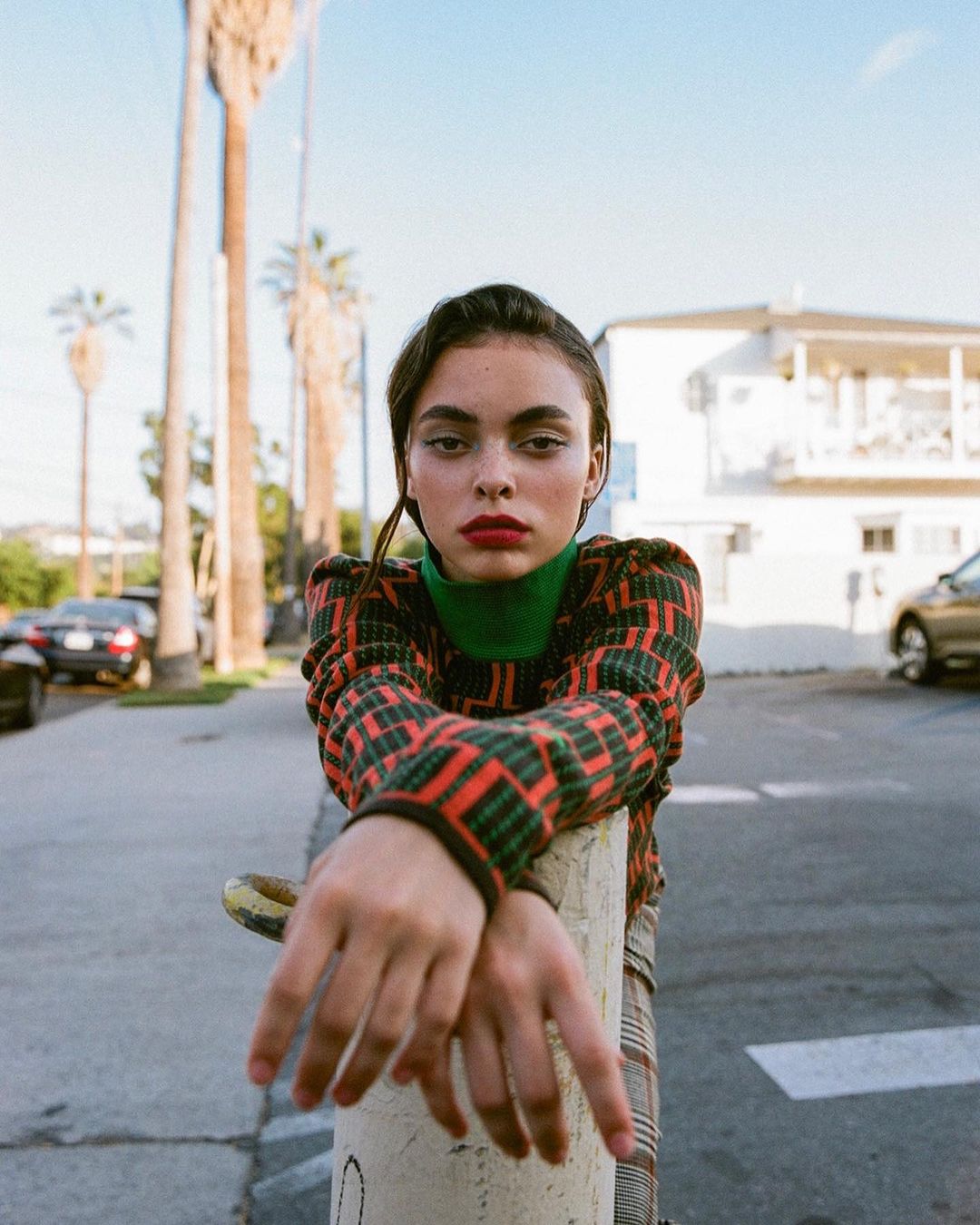 Young woman in patterned sweater leans against pole in urban setting with palm trees and cars in the background on sunny day.