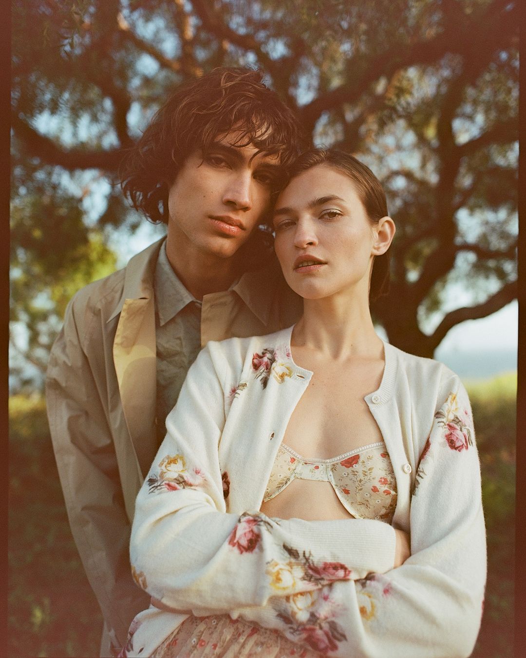 A stylish couple poses outdoors in a sun-drenched shot with visible bra and a light floral cardigan, under a tree canopy.
