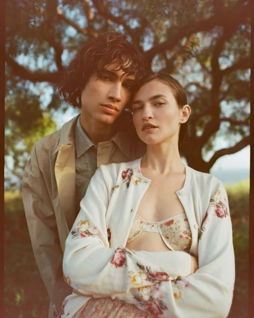 A stylish couple poses outdoors in a sun-drenched shot with visible bra and a light floral cardigan, under a tree canopy.