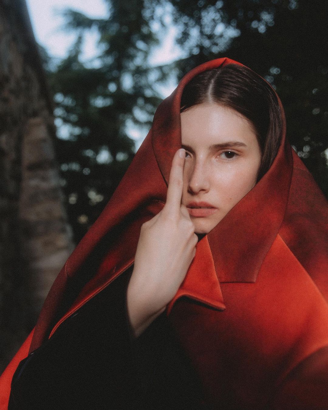 Woman with a red fabric draped over her head, partially covering her face, posing outdoors against a backdrop of blurred trees.