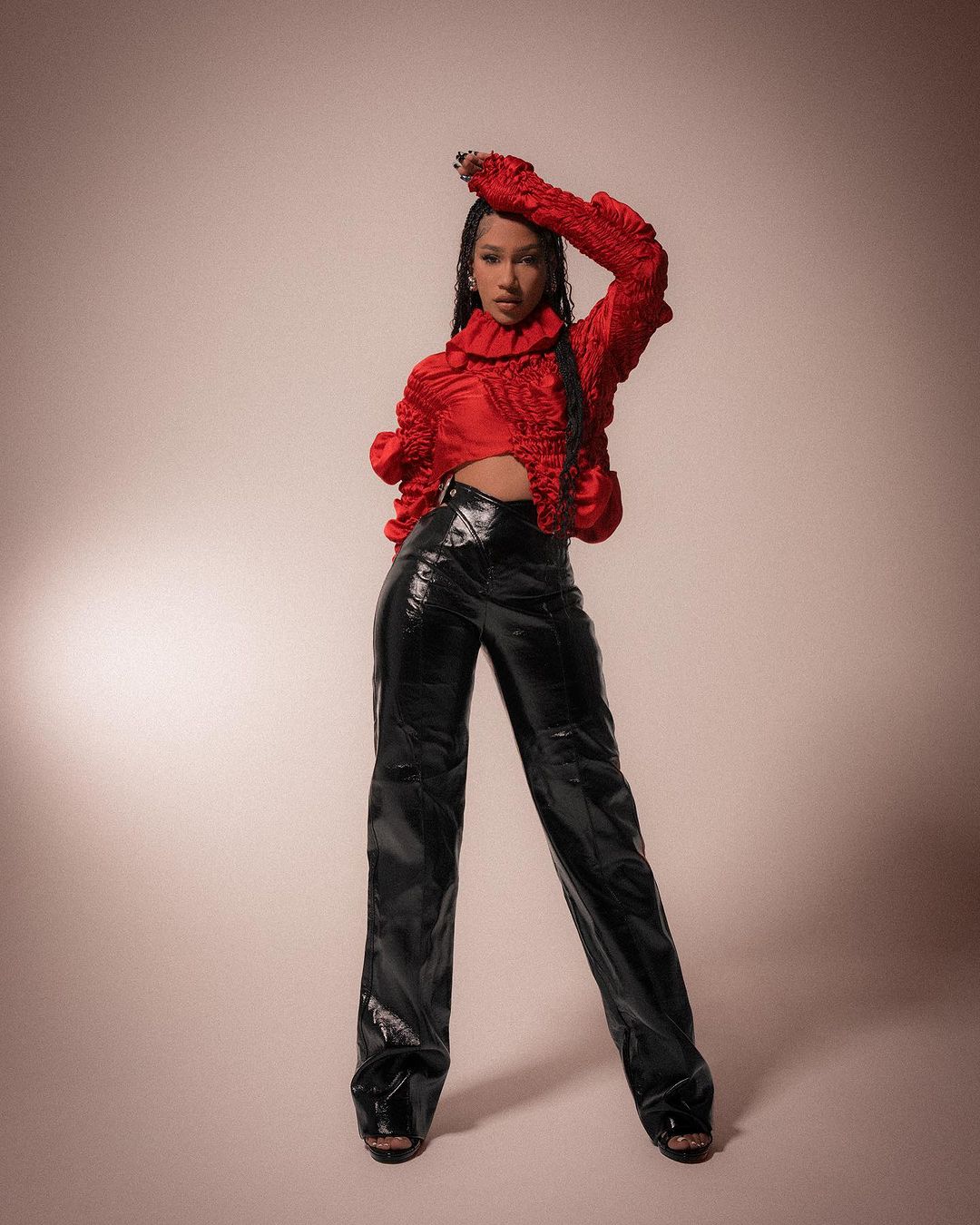 Full-length portrait of an adult female in a red top and shiny black pants posing in front of a neutral-toned backdrop in studio light.