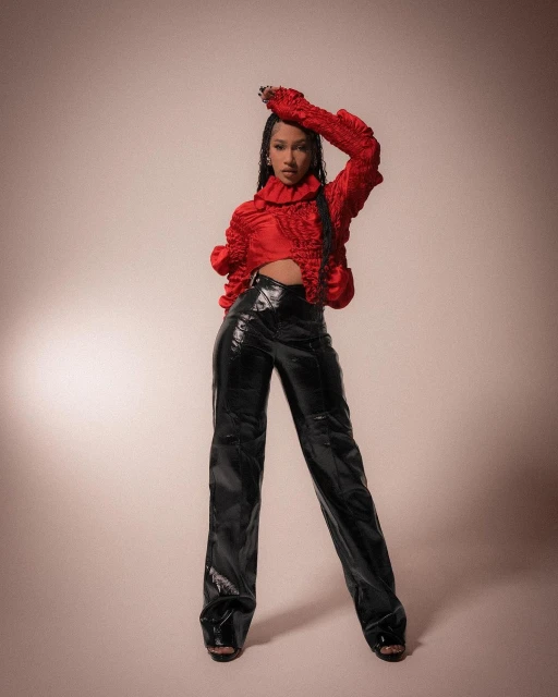 Full-length portrait of an adult female in a red top and shiny black pants posing in front of a neutral-toned backdrop in studio light.