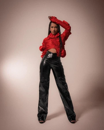 Full-length portrait of an adult female in a red top and shiny black pants posing in front of a neutral-toned backdrop in studio light.