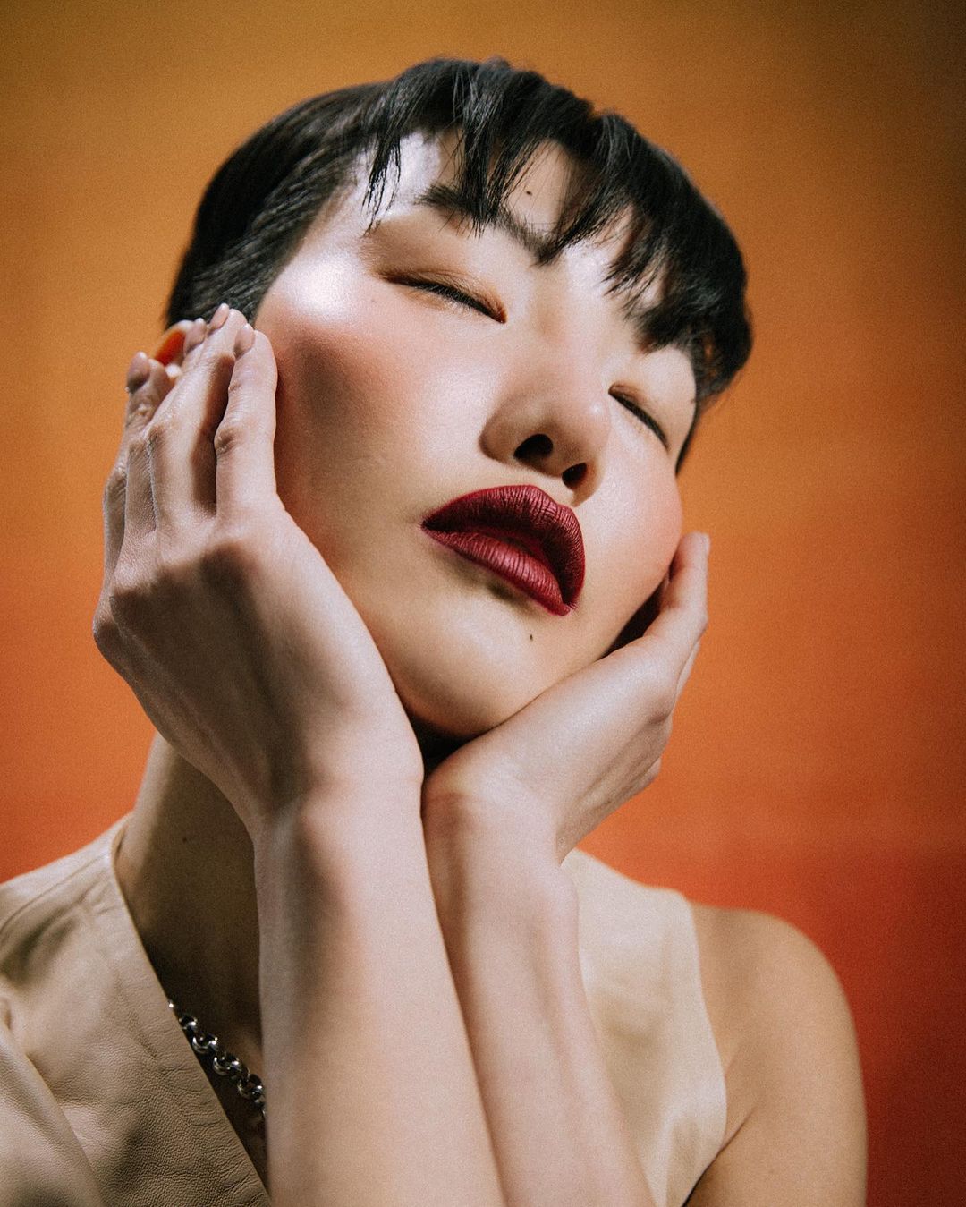 Close-up portrait of an Asian woman with dark lipstick and closed eyes, softly holding her face against a gradient orange background.
