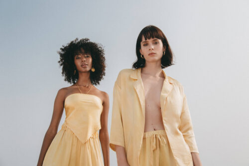 Two women in light yellow linen outfits stand against a clear sky, one in a strapless top and the other in an open blazer.