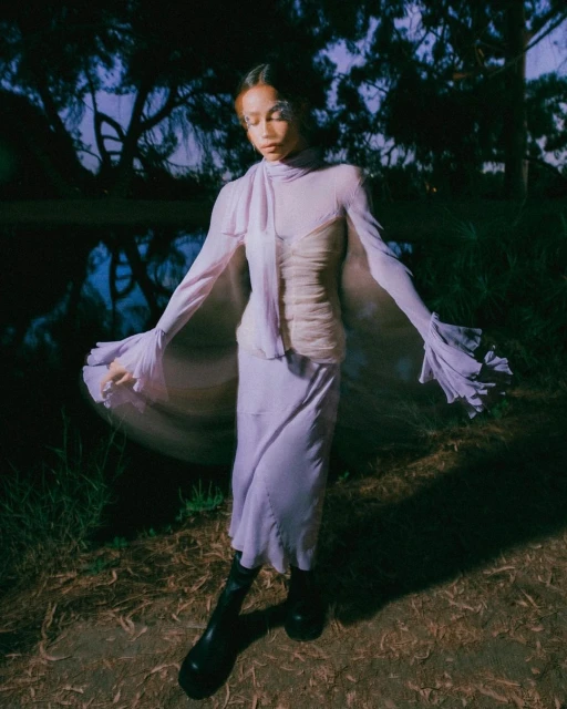 Woman in flowing lilac gown poses near pond surrounded by trees in dusk light, exuding a serene fashion editorial vibe.