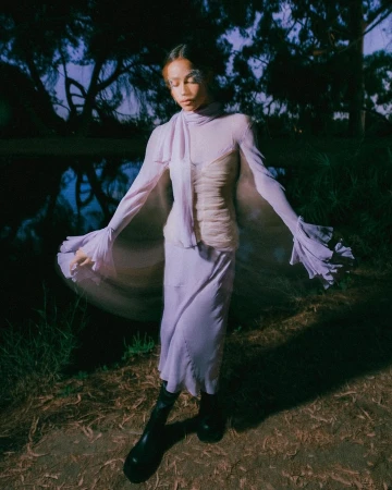 Woman in flowing lilac gown poses near pond surrounded by trees in dusk light, exuding a serene fashion editorial vibe.