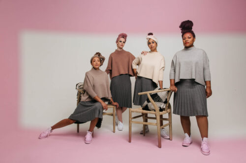 Group portrait of four women in matching gray skirts and neutral tops, wearing headscarves, posing in a photo studio with pink backdrop.