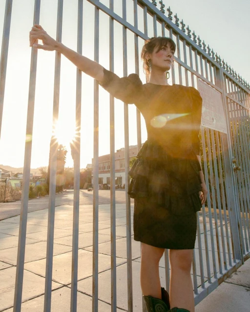 An adult woman stands behind a metal gate in a dress with ruffles and cowboy boots as the sun shines behind her.